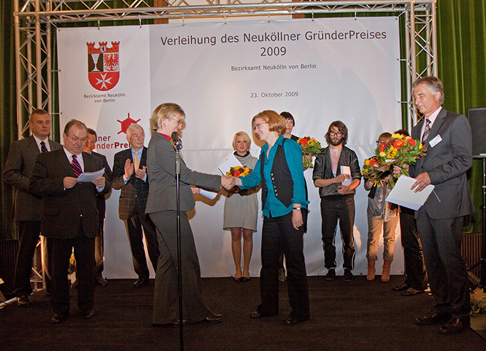 Foto von der Preisverleihung beim Neuköllner Gründerpreis am 23. Oktober 2009 im Bezirksamt Neukölln von Berlin, auf der Bühne wird Nicola Pridik gerade von einem Jurymitglied gratuliert.